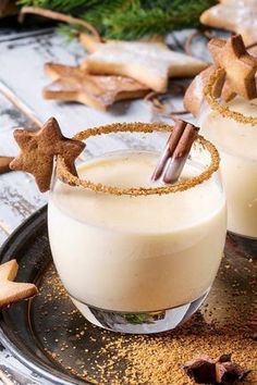 two glasses filled with drink sitting on top of a metal tray next to cookies and cinnamon sticks