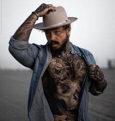 a man with tattoos on his chest wearing a cowboy hat and denim jacket standing in front of the ocean