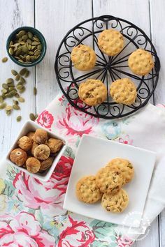 some cookies are on a white plate and two bowls with nuts in them next to a flowered towel