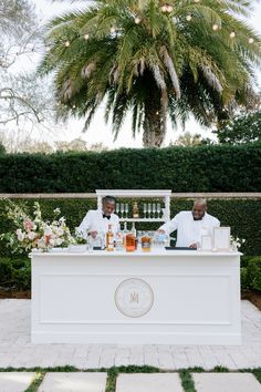 two men sitting at a white bar with drinks on it in front of a palm tree