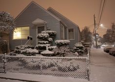 a house covered in snow at night with cars parked on the street and behind it is a chain link fence