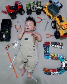 a baby laying on the ground surrounded by toy cars and tools, including a truck