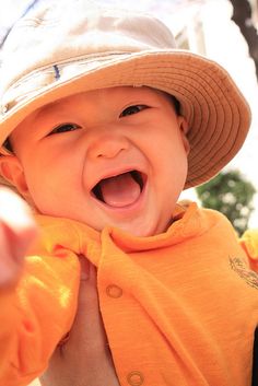 a baby wearing an orange bib and a tan hat smiles as he holds his hand out