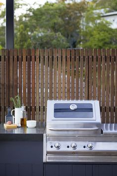 an outdoor kitchen with a grill, sink and coffee mug on the counter top in front of a wooden fence