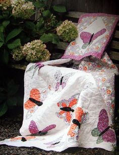 a quilted blanket and pillow on a bench next to some flowers in the background