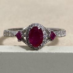 an oval shaped ruby and diamond ring sits on top of a white stone block in front of a beige background