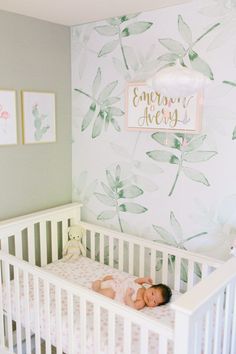 a baby laying in a white crib next to a wall with green leaves on it