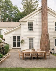 a table and chairs in front of a white house with a brick chimney next to it