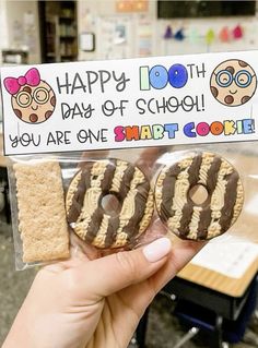 a person holding up two cookies in front of a sign that says happy 100th day of school you are one smart cookie