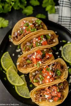 four tacos on a black plate with limes and cilantro wedges