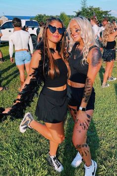 two women in black outfits posing for the camera at an outdoor music festival with other people