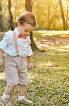 a young boy wearing a bow tie and suspenders in the grass with trees behind him