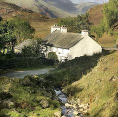 a small white house sitting on the side of a hill next to a stream and trees