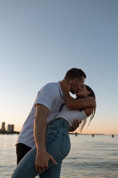 a man and woman kissing in front of the ocean at sunset, with buildings in the background