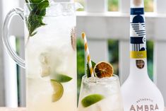 two glasses filled with lemonade and limes next to a bottle of ambra