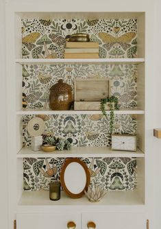 an open bookcase with books and other items on it, in front of a wallpapered background