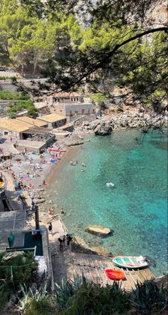 the beach is crowded with people and boats in clear blue water, surrounded by pine trees