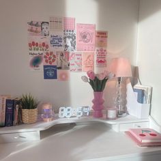 a white desk topped with a pink vase filled with flowers next to a wall covered in pictures