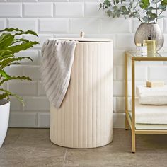 a white laundry basket next to a potted plant and towel on a stand in front of a brick wall