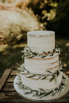 a three tiered cake with greenery on top sits on a wooden table outside