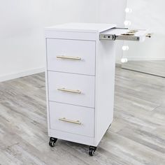 a white filing cabinet with three drawers on casteors and two lights in the background