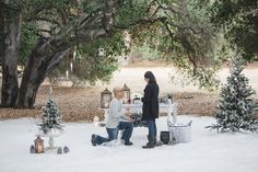 two people are sitting in the snow near christmas trees and lanterns, one person is holding a lantern