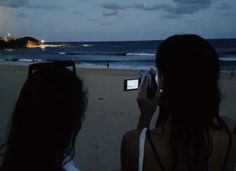 two women are taking pictures on the beach with their cell phones at night, and one woman is holding up her camera