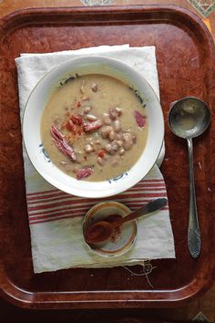 a bowl of soup with beans and ham on a wooden tray next to a spoon