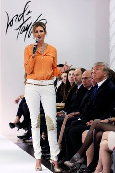 a woman in an orange shirt and white pants is standing on the runway with a microphone