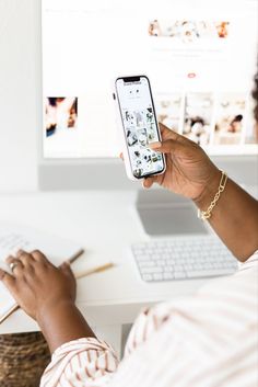 a woman holding up her cell phone while sitting at a desk with a computer and keyboard