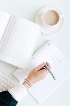 a person writing on a notepad next to a cup of coffee and an open book