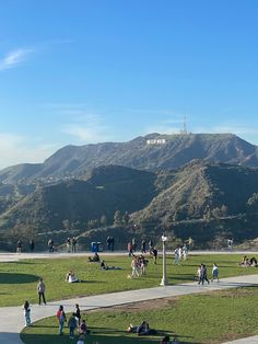 many people are walking and sitting on the grass in front of some hills with a hill in the background