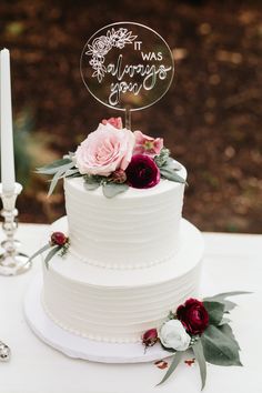 a white wedding cake with pink flowers and greenery sits on a table next to two candles