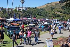 a crowd of people walking down a street next to parked cars and tents in the grass