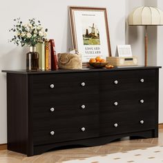 a black dresser with drawers and pictures on the top, next to a lamp in a living room