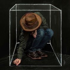 a man sitting in a glass box with his head down wearing a hat and jeans