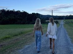 two women walking down a dirt road in the middle of a grassy field at dusk