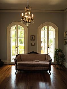 a couch sitting in the middle of a living room next to two large windows and a chandelier