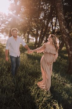 a couple holding hands and walking through the grass in front of some trees at sunset