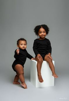 two young children sitting on top of a white block posing for the camera with their hands in each other's pockets