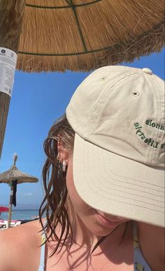 a woman with long hair wearing a hat and sitting under an umbrella on the beach