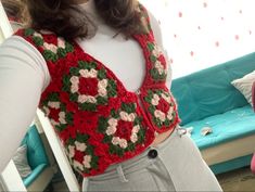 a woman standing in front of a couch wearing a red crocheted vest with flowers on it