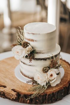 a three tiered cake with white flowers and greenery sits on a wooden slice