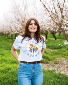 woman modeling and standing in a blooming almond blossom orchard wearing upper park's california grown cropped tee in white paired with light wash denim jeans and a brown leather belt. the shirt depicts orange california poppies with green text reading "california grown". almond orchard photoshoot, flowers, almond season, california, modeling photography Spring Graphic Tee Crop Top, Cotton Graphic Tee Crop Top, Strawberry Print Crew Neck Graphic Tee, Fitted Cotton Graphic Tee Cropped T-shirt, California T Shirt Designs, Hot Desert, We Are All Human, California Poppy, Snowy Mountains