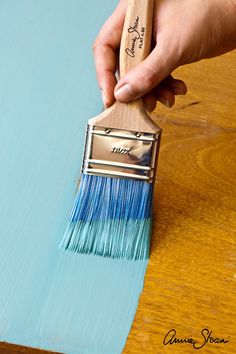 a person holding a paint brush on top of a wooden table next to a blue wall