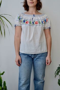 a woman standing next to a potted plant wearing jeans and a white top with flowers on it