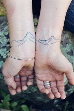 two people holding hands with tattoos on their wrists and mountains in the background, both showing