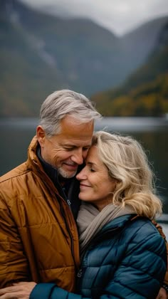 an older couple embracing each other in front of mountains and the water on a cloudy day