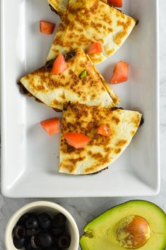 three quesadillas on a white plate with an avocado and black olives