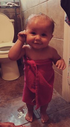 a baby in a red towel is standing next to a toilet and looking at the camera